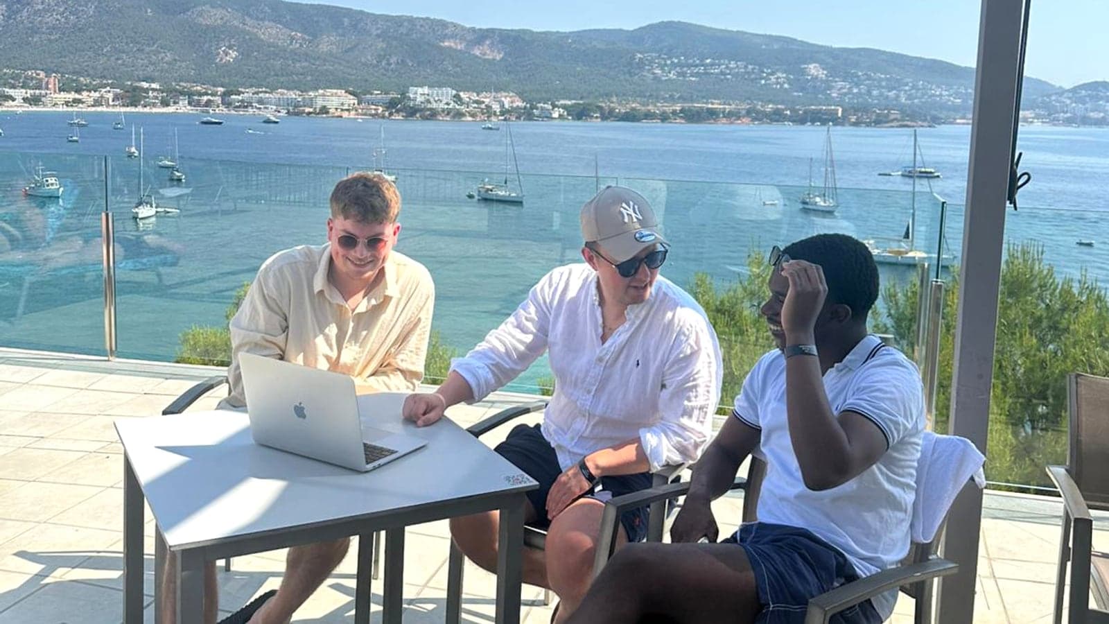 Drei Mitglieder der Zuversichtsberatung sitzen während einer Workation auf einer Terrasse über der Bucht von Palmanova auf Mallorca am Laptop